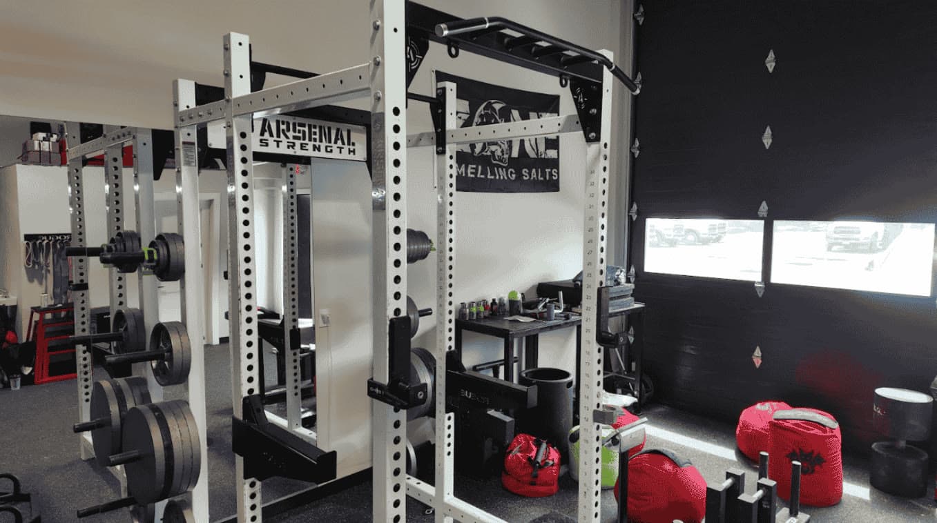 White Arsenal Strength power racks with multi-grip pull-up bars beside strongman sandbags and an open garage bay door