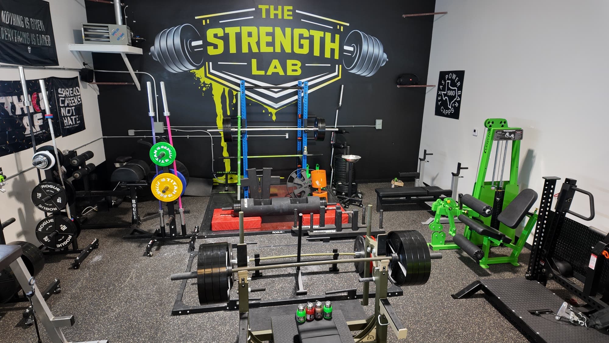 The Strength Lab floor in Newtown, CT: the black and green logo mural above a blue power rack, a loaded deadlift bar, calibrated plates, strongman logs on the platform and the green Legend Fitness machines along the wall
